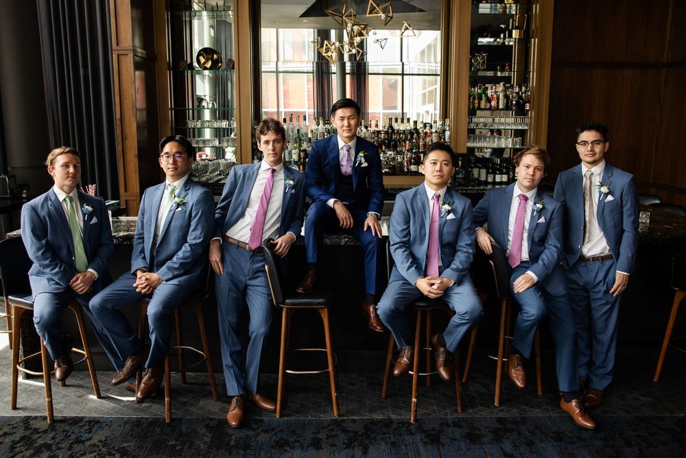 Seven groomsmen in blue suits with pink ties pose confidently at a stylish bar venue for a wedding photo.