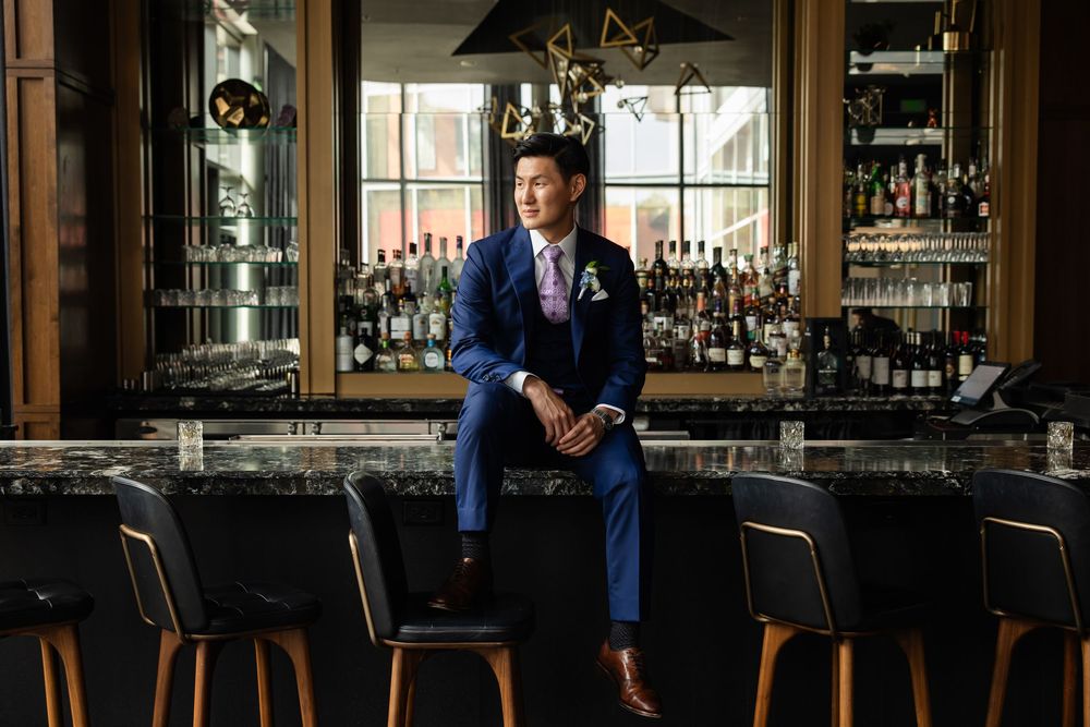 Man in blue suit sitting at upscale bar with marble countertop and liquor bottles in background.