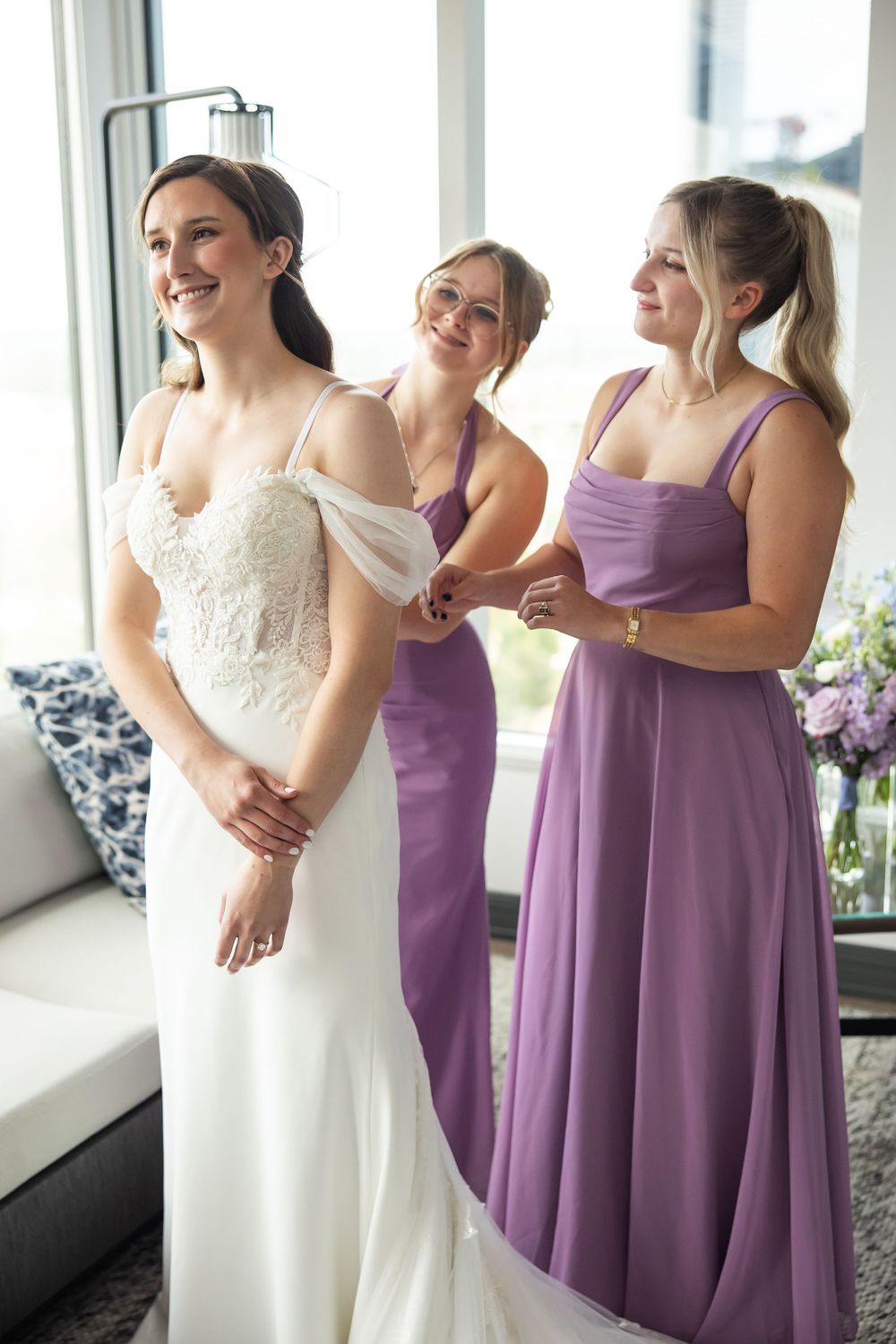 Bride smiling in white lace gown while two bridesmaids in purple dresses help her get ready indoors.