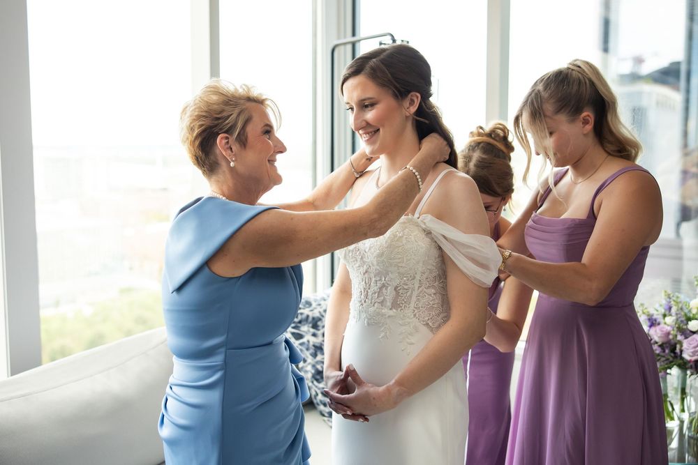 Bride being helped into wedding dress by mother and bridesmaid in purple gown near large windows.