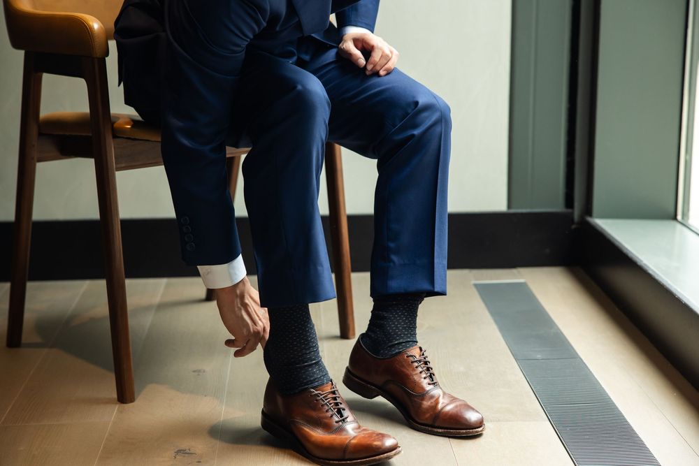 Man in navy blue suit adjusting brown leather oxford shoes while seated, wearing dark dress socks.