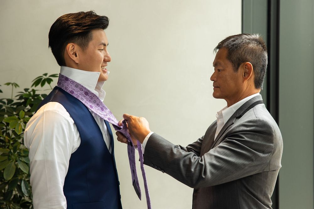 An older man helps a young man tie a purple necktie over a white dress shirt and blue vest.