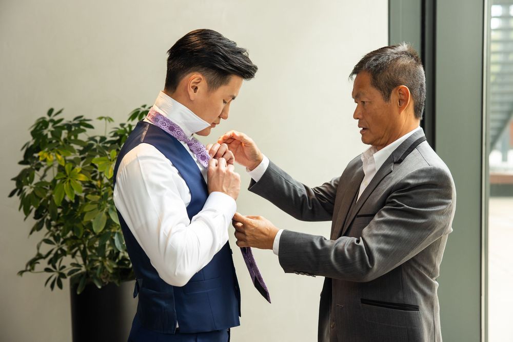 Two men in formal suits helping each other adjust ties and cufflinks before a wedding ceremony.