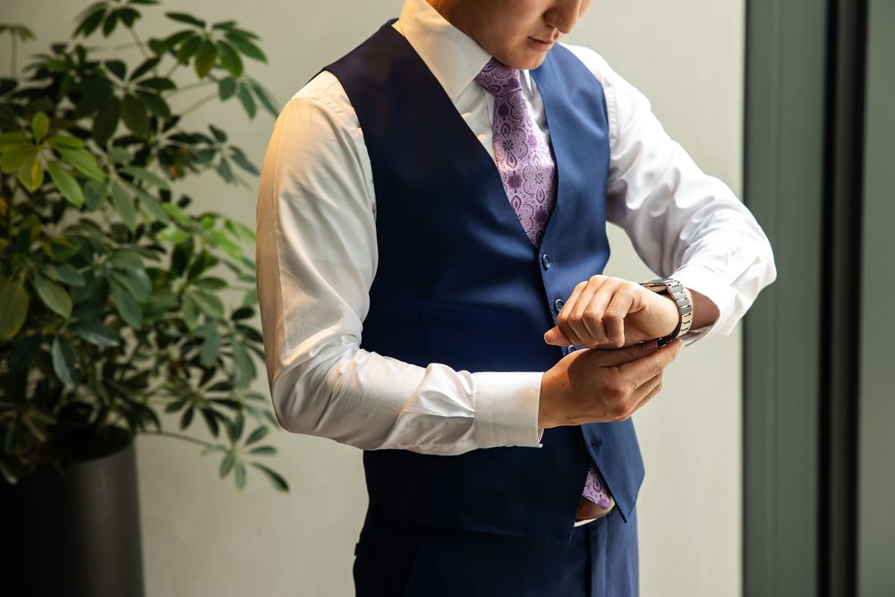 Businessman in blue vest and tie checking his watch while holding a smartphone in an office setting.