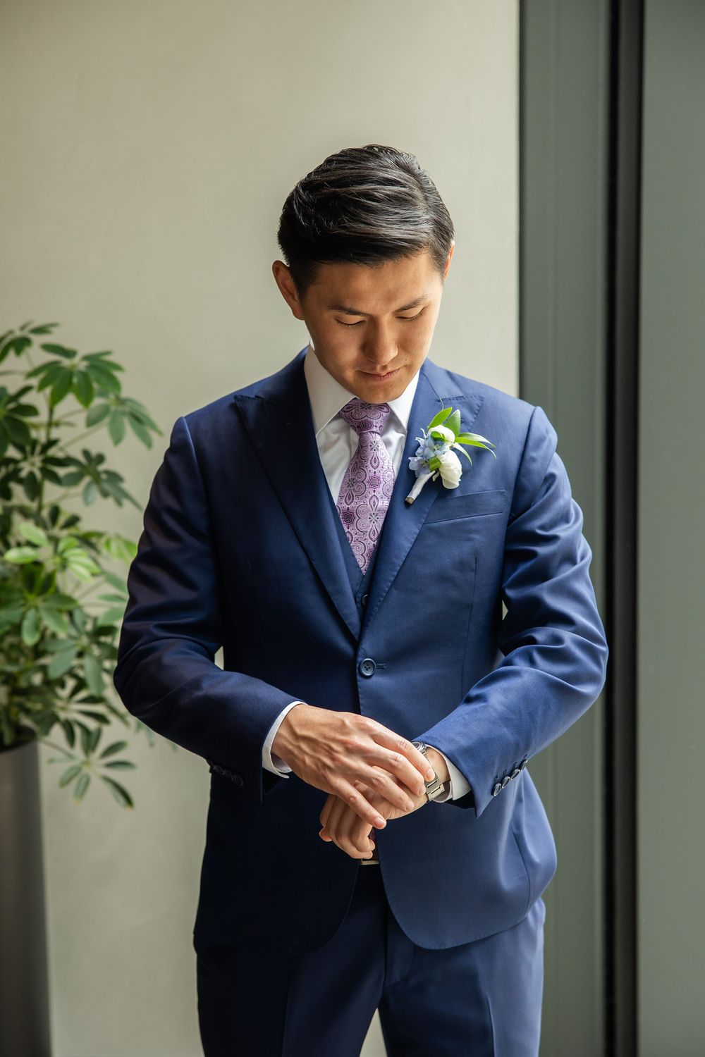 Groom in navy blue suit with floral boutonniere and purple tie adjusting cufflinks before wedding ceremony.