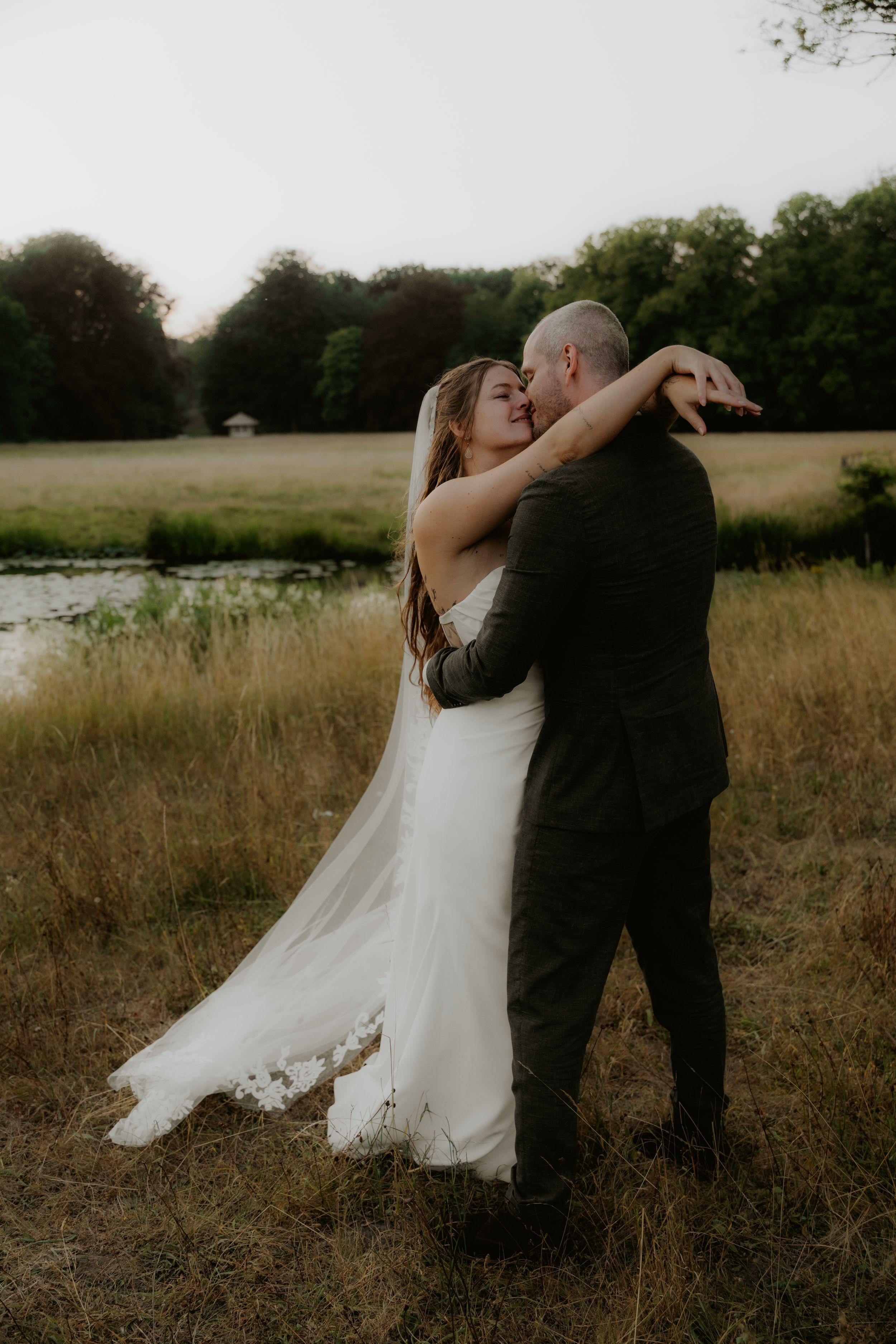 A bride and groom share a romantic embrace in a golden field at dusk, with a serene pond and lush trees in the background.