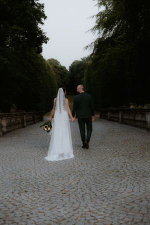 Bride and groom walking hand in hand down a cobblestone path lined with tall green trees on their wedding day.