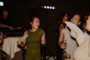 Women dancing joyfully at a wedding reception, holding fans and drinks, with a DJ setup visible in the background.