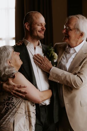 A groom in a suit with boutonniere shares a warm moment with his smiling elderly parents on his wedding day.