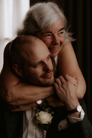 Older woman with gray hair hugging a bald groom in a suit with a white rose boutonniere at a wedding.