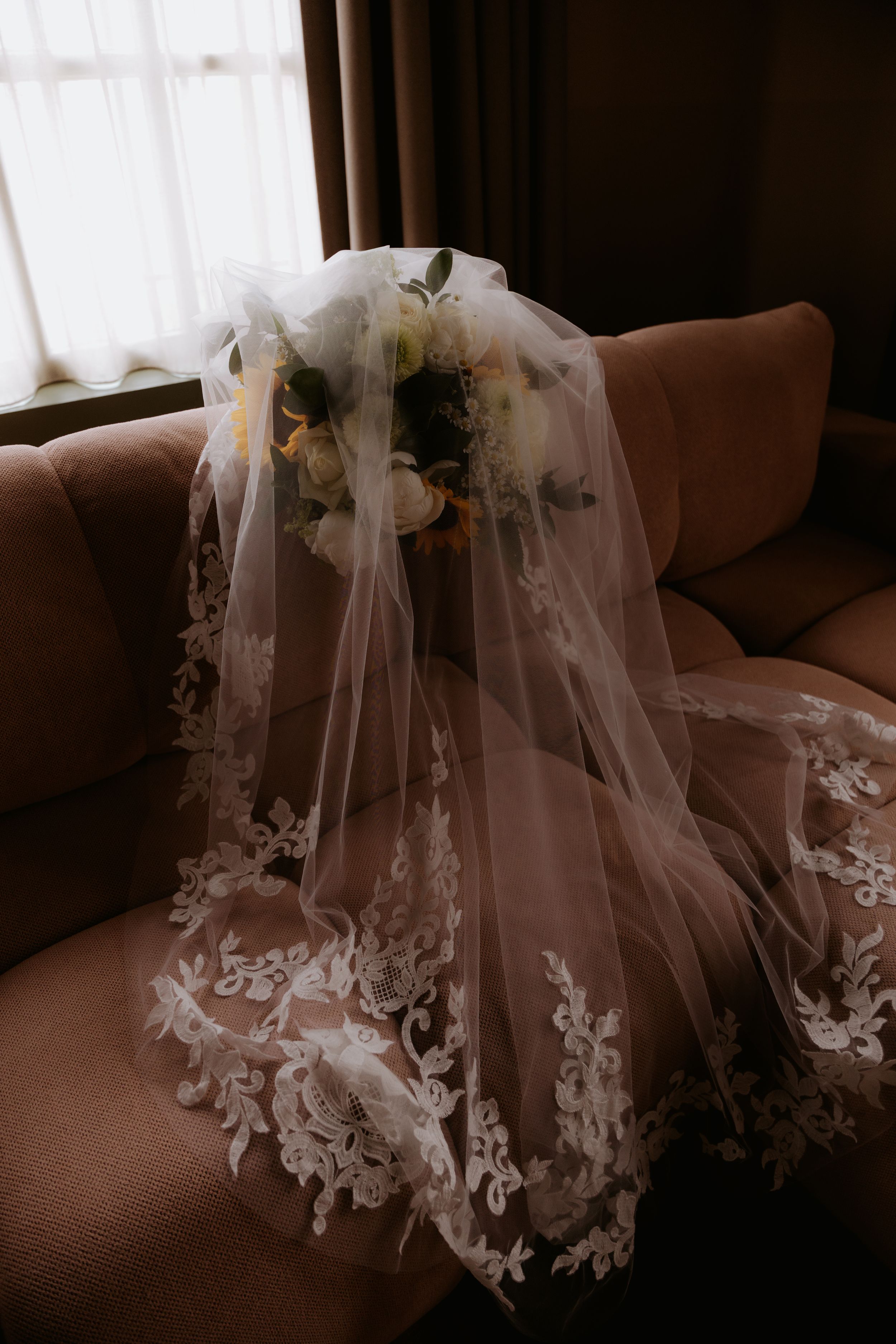 Bridal veil with lace trim draped over a bouquet of white and yellow flowers on a pink sofa near a window.
