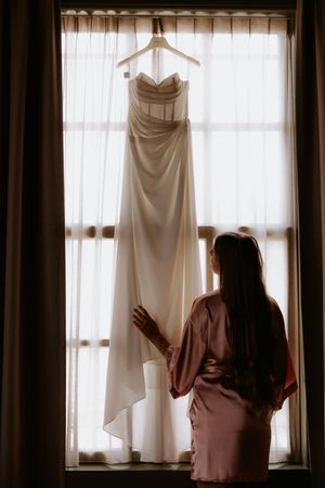 Bride in silk robe admiring her elegant strapless wedding gown hanging by a sunlit window.