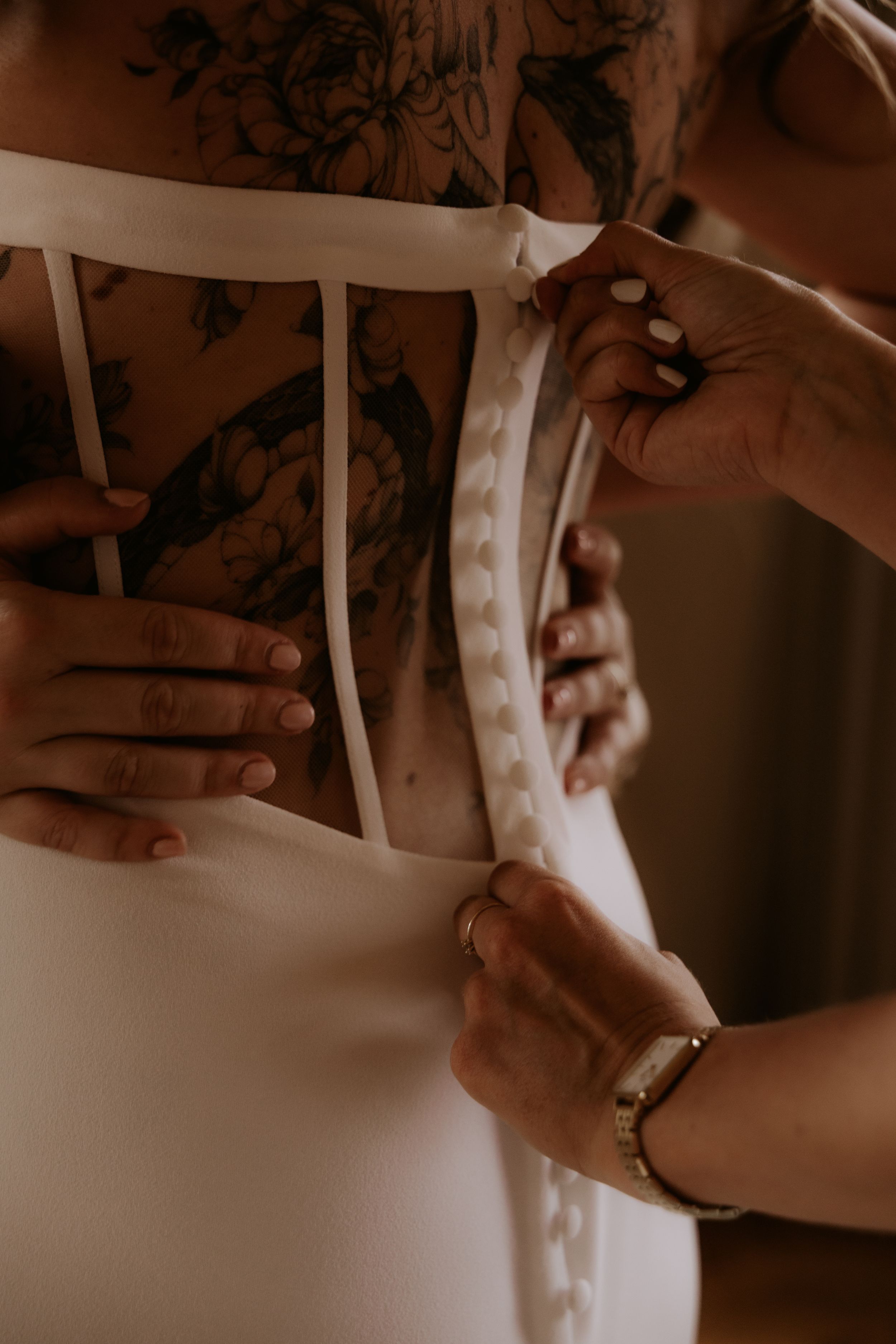 Tattooed bride getting her white wedding dress buttoned up the back by a helper with manicured nails.
