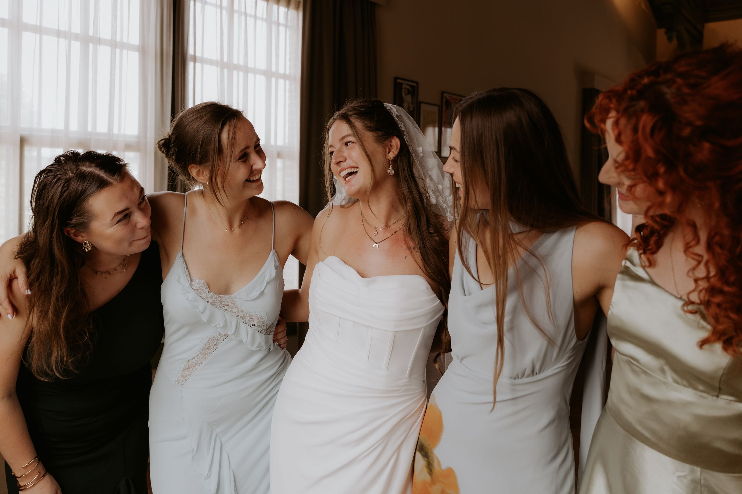 Bride in white strapless gown laughing joyfully with bridesmaids in silver dresses during wedding preparations.