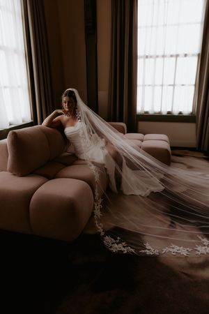 Bride in strapless gown with long lace-trimmed cathedral veil seated on blush sofa in elegant room.