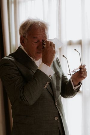 Elderly man in gray suit holding glasses while wiping his eye with a tissue near a bright window.