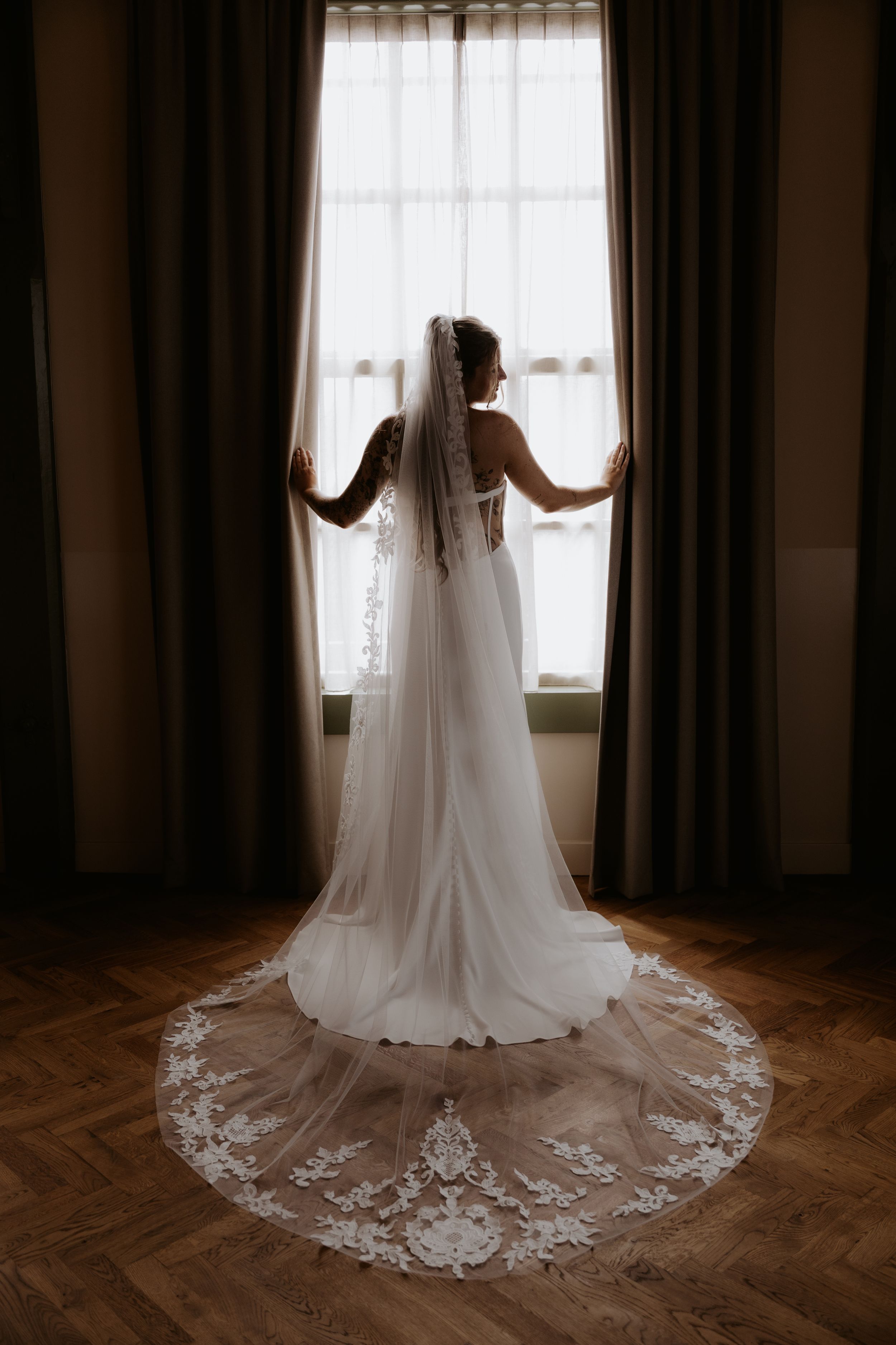 Bride in white lace wedding gown and cathedral veil stands before a sunlit window in elegant venue.