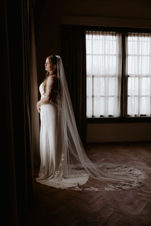 Bride in a white lace long-sleeve gown and cathedral veil stands by a window in a dimly lit room.