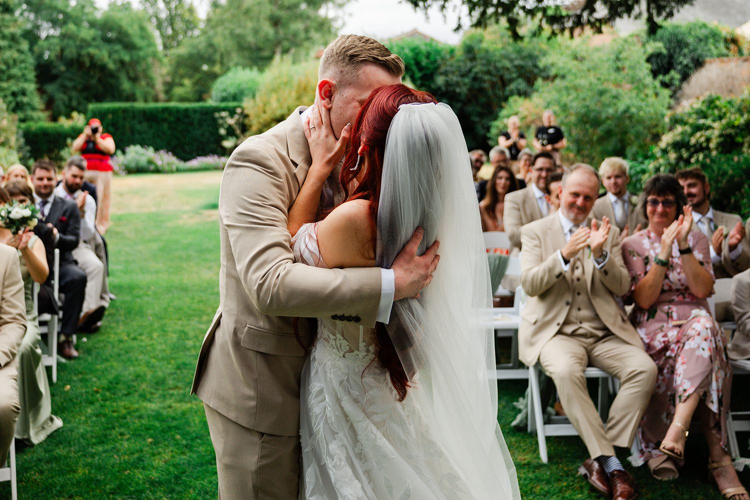 A groom in a tan suit kisses his bride in a white lace gown and veil at an outdoor garden wedding ceremony.