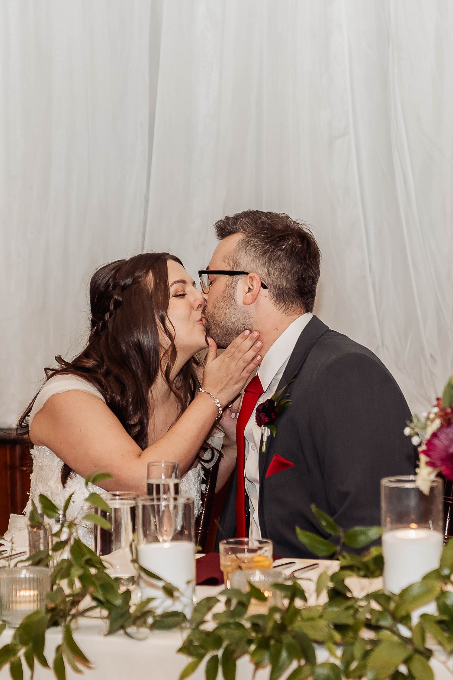 A bride and groom share a romantic kiss at their wedding reception table adorned with greenery and candles.