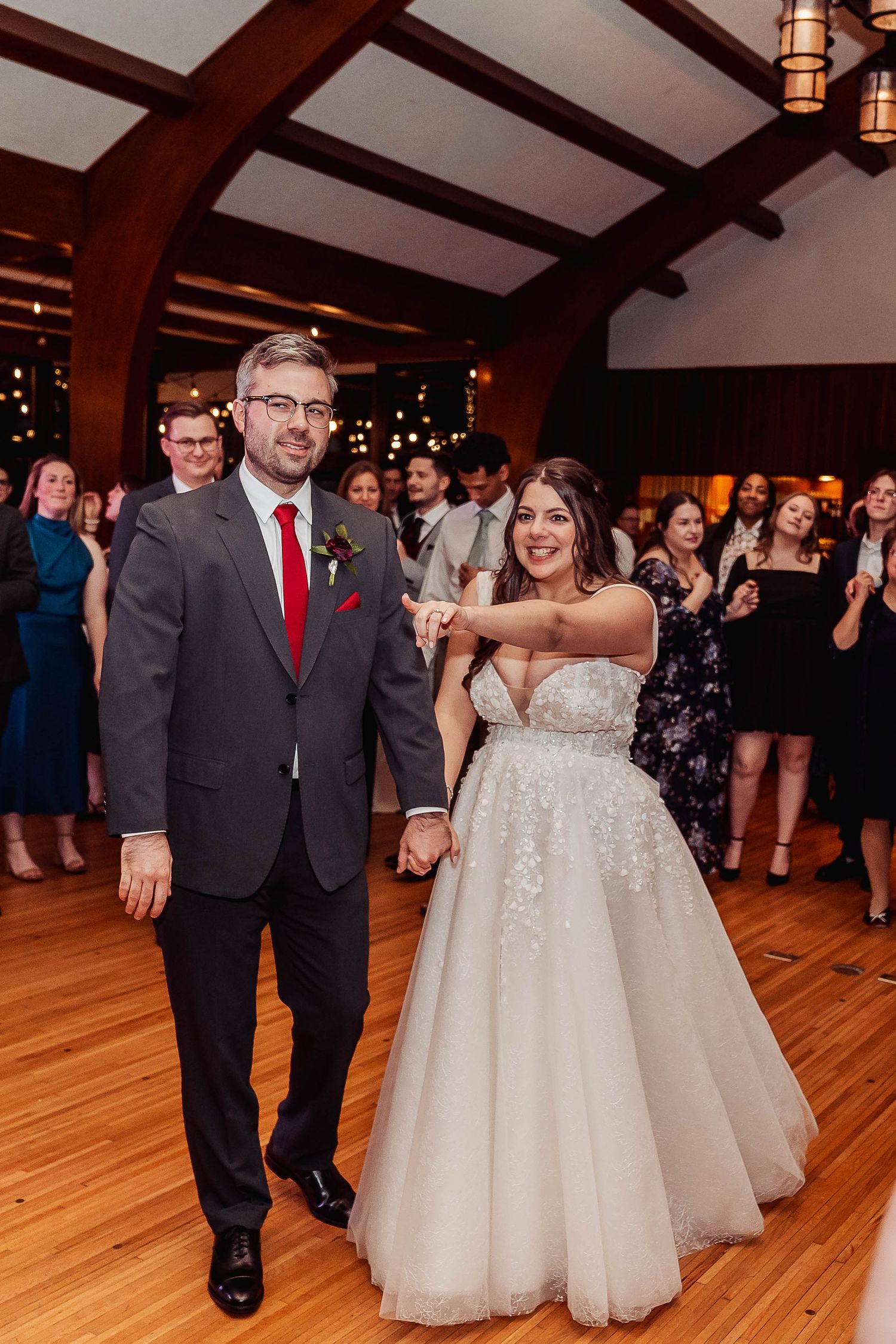 A bride and groom share their first dance at a wedding reception in an elegant wood-beamed venue with guests watching.