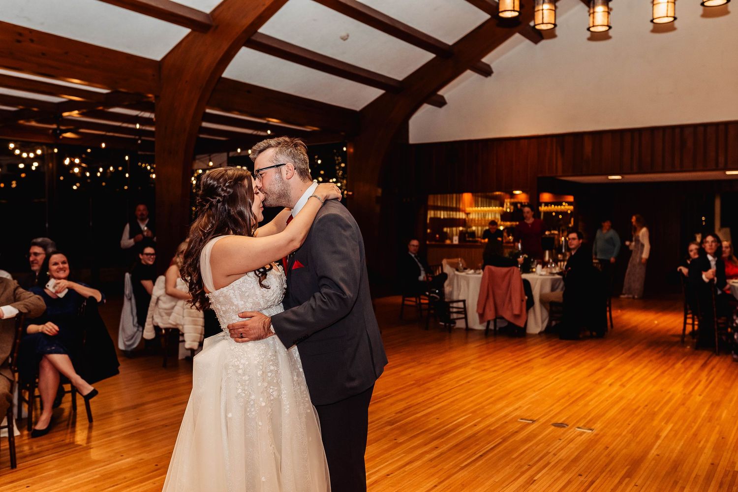 Bride and groom share their first dance on a wooden floor in a rustic wedding venue with warm string lights.