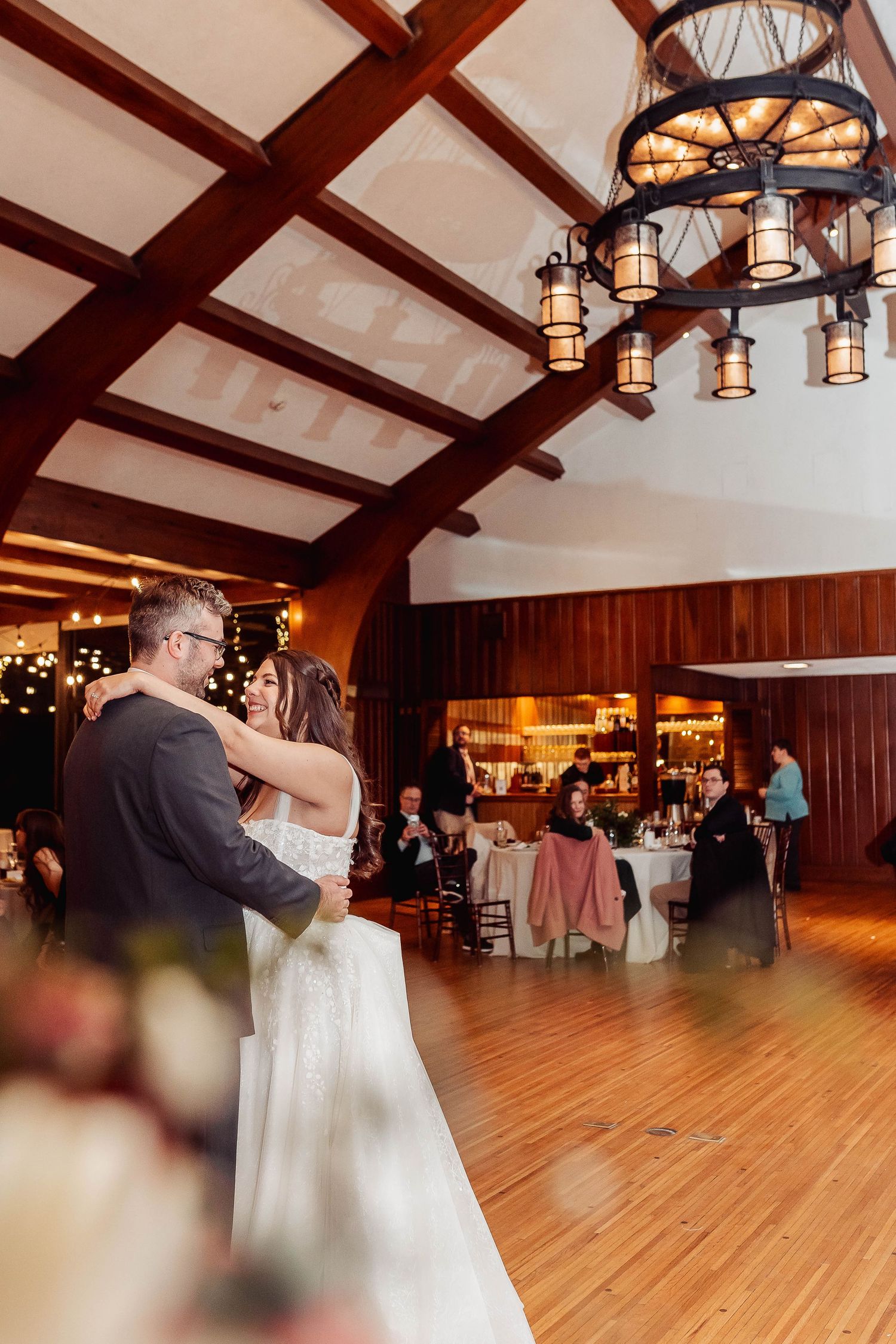 Bride and groom share their first dance on a wooden floor in a rustic venue with exposed beams and chandelier.