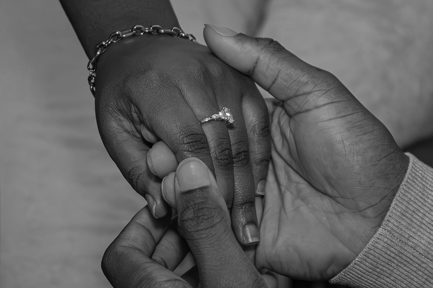 Black and white photo of two hands holding, one wearing an engagement ring and bracelet, symbolizing love and commitment. Engagement