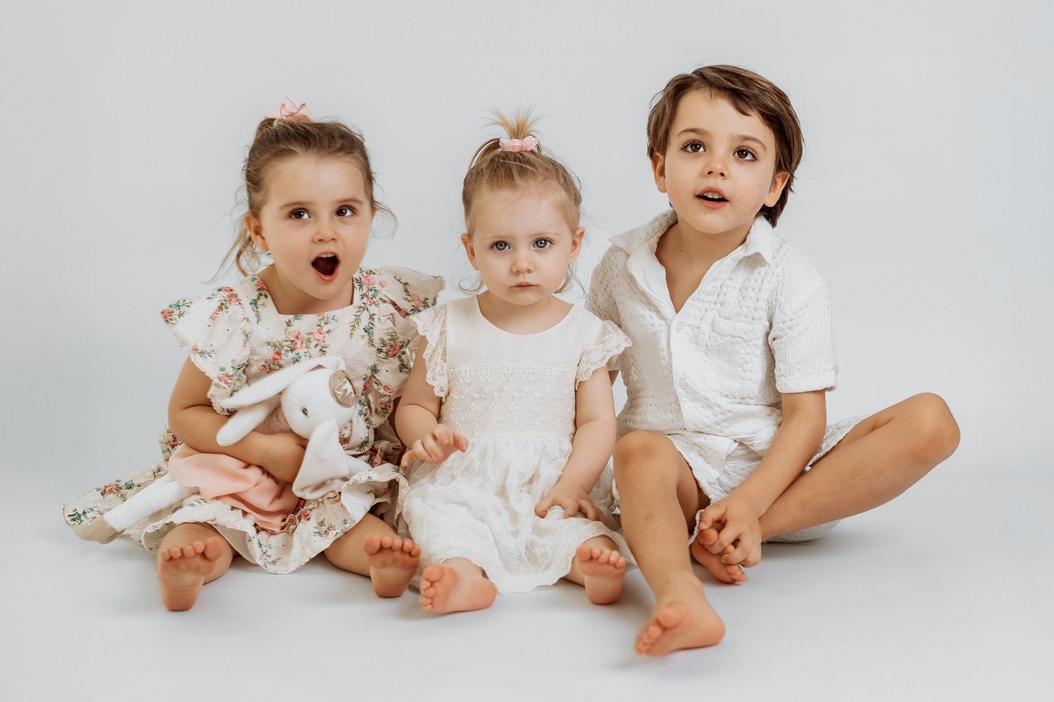 Three children in matching soft neutral outfits sitting on a white studio backdrop with a plush bunny in Dubai
