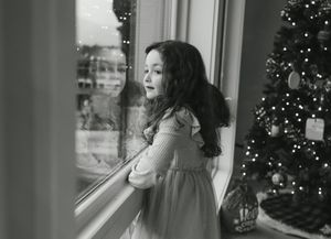Black and white photo of a young girl gazing out a window beside a decorated Christmas tree.