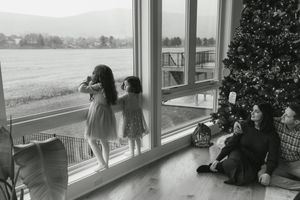 Two girls look out large windows at a snowy landscape beside a Christmas tree while a woman sits on the floor nearby.