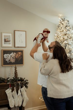 A couple lifts their baby girl near a decorated Christmas tree with stockings and framed art in a cozy holiday home.