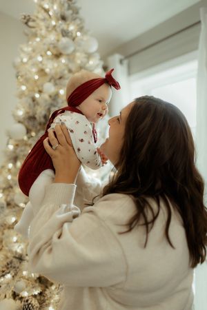 Mother holding baby girl in red bow near lit Christmas tree, both in festive white and red outfits.