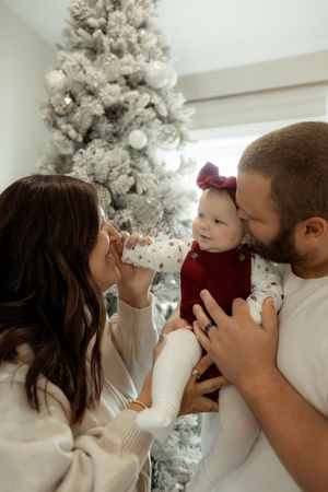 A couple shares a joyful Christmas moment with their baby girl in front of a flocked white holiday tree.