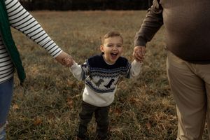 A joyful toddler laughs while being held by both hands by two adults in a grassy autumn field.