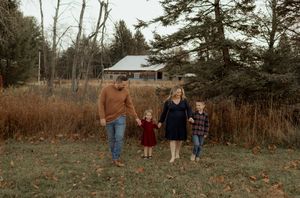 Family of four walking hand-in-hand through a golden autumn field with evergreen trees and a red barn in the background.