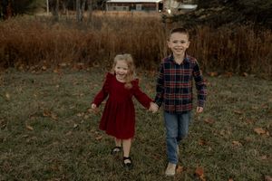 Two young children holding hands and walking through a grassy field in autumn, wearing red and plaid outfits.