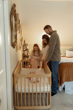 Family of three admiring newborn baby in wooden crib in a warmly lit nursery bedroom.