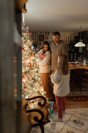 Family of four admiring a decorated Christmas tree with glowing lights in a cozy, dimly lit living room.