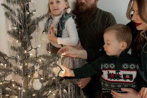 Family decorating a Christmas tree together with fairy lights, parents and two young children in festive sweaters.