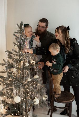 Family of four decorating a Christmas tree with white ornaments and fairy lights in a bright white studio.