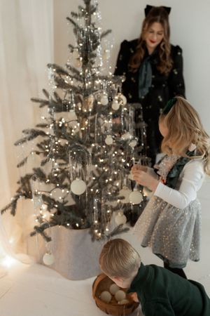 Young girl and boy decorating a silver Christmas tree with white ornaments while mother watches nearby.