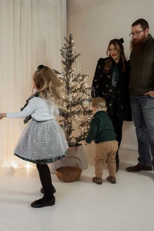 Family with two young children playing near a lit Christmas tree in a bright white holiday photo studio.