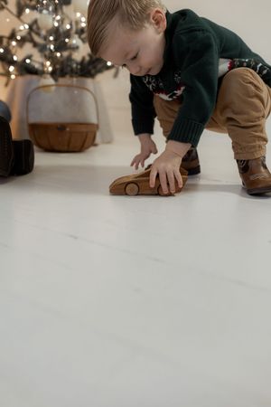 Toddler in green sweater playing with wooden toy car on white floor near Christmas tree with lights.