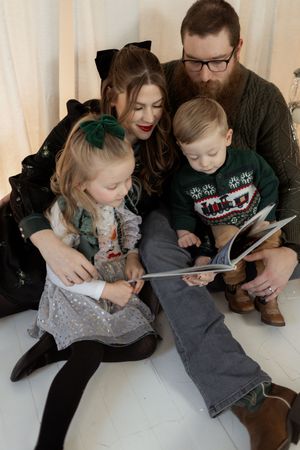 Family of four sitting together reading a book, wearing festive holiday outfits including ugly Christmas sweater.