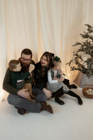 Family of four sitting together by a decorated Christmas tree, smiling and enjoying the holiday season indoors.