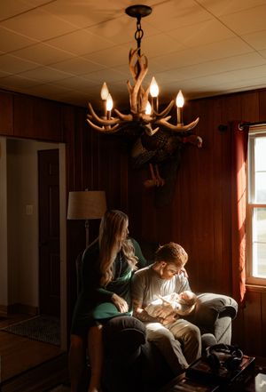 Family with newborn baby sitting in armchair under antler chandelier in rustic wood-paneled room with moose mount.