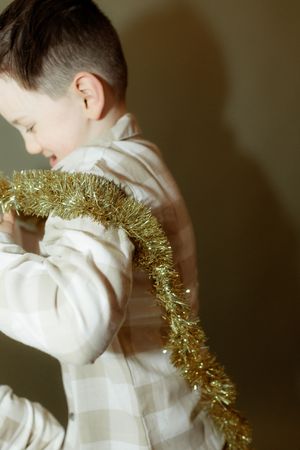 Young boy in plaid pajamas holding gold tinsel garland, holiday Christmas decoration activity.