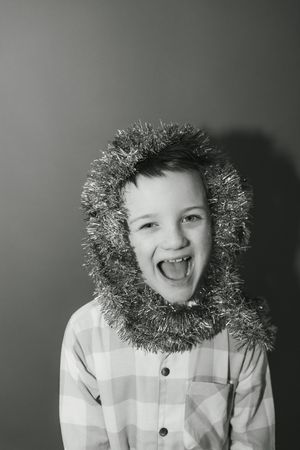 Black and white photo of a laughing boy wearing Christmas tinsel garland as a hood over his plaid shirt.