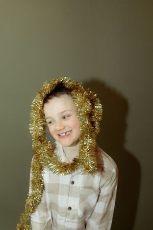 A smiling child wearing a plaid shirt adorned with gold Christmas tinsel draped over their head and shoulders.